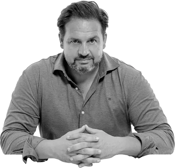 Hombre de mediana edad con barba y cabello oscuro, vestido con una camisa gris, sentado con las manos entrelazadas y una expresión seria.